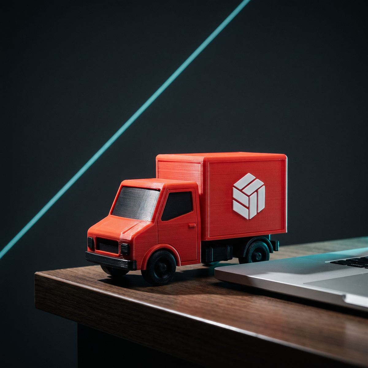Miniatura de caminhão de entrega vermelho impressa em 3D sobre uma mesa, ao lado de um notebook